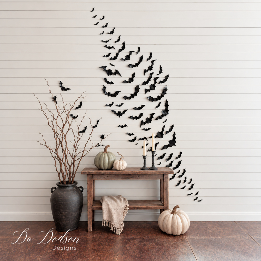 Black bats arranged in a sweeping Halloween bat wall above a rustic console table decorated with neutral pumpkins, candles, and bare branches.