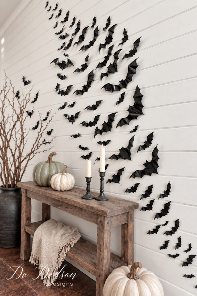 Black three-dimensional bats sweeping across a white shiplap wall above a rustic console table styled with neutral pumpkins and black candlesticks.