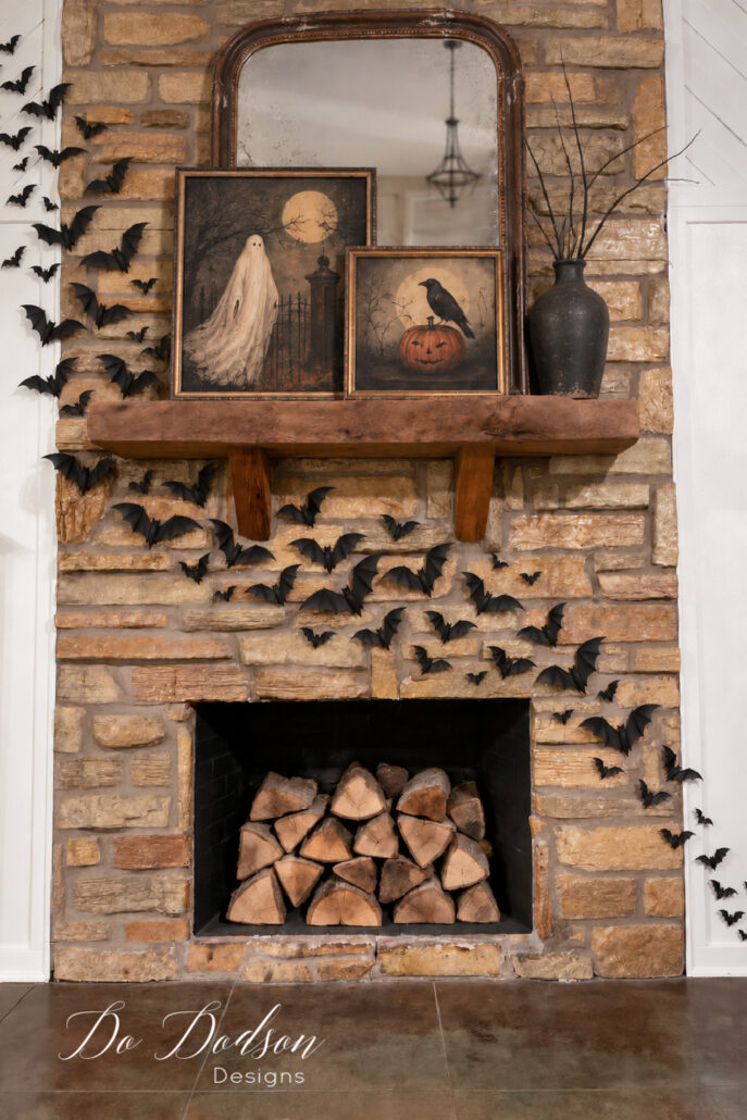 Black 3D PVC bats arranged across a rustic stone fireplace with stacked firewood, vintage Halloween artwork, an antique mirror, and a black vase with bare branches.
