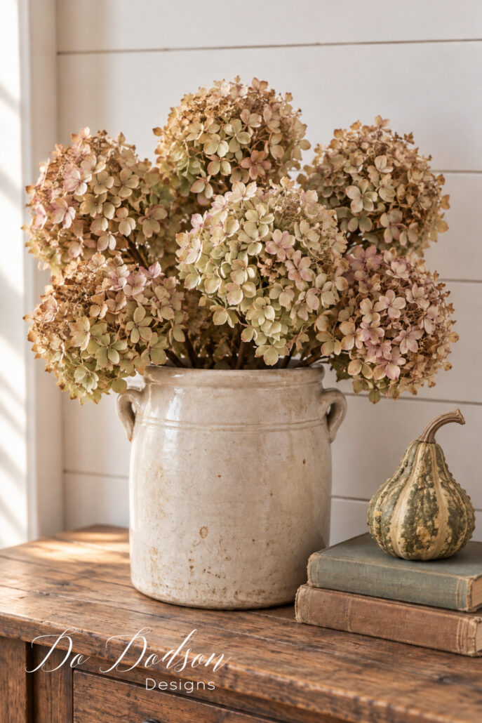 Dried green and blush hydrangeas in a vintage crock styled with a natural fall gourd and old books