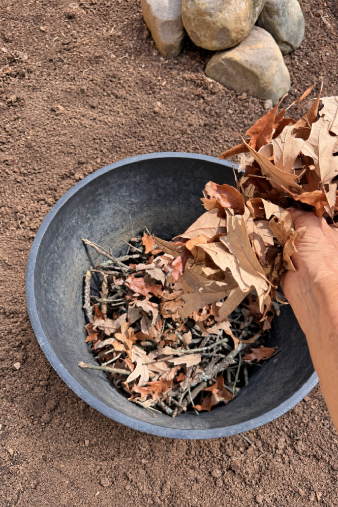 Hand adding dry fallen leaves over small sticks in the bottom of a large black planter.