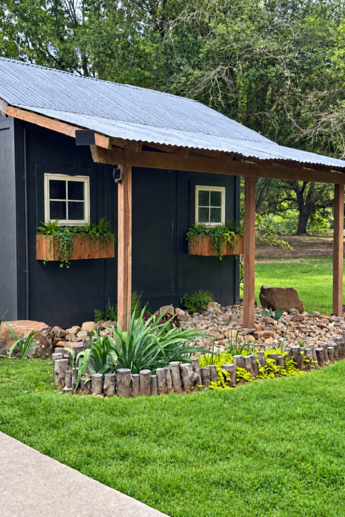 Modern farmhouse shed painted Sherwin-Williams Iron Ore with stained cedar window boxes, natural greenery, and a low-maintenance river rock garden.