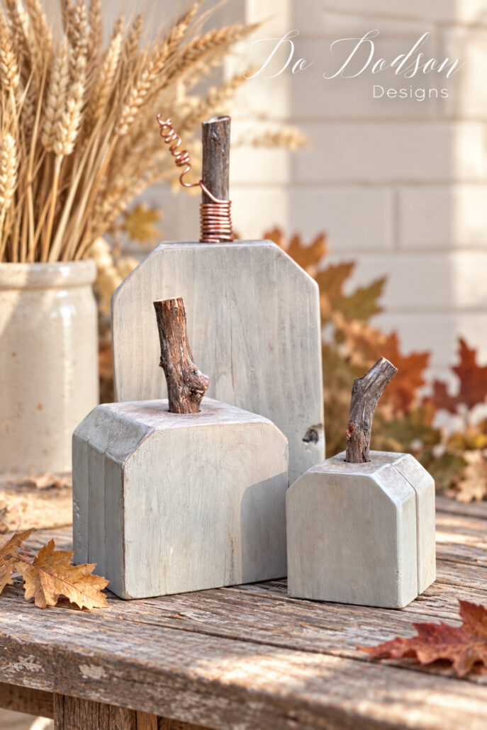 Three handmade white wood pumpkins with natural branch stems and a copper wire curl displayed on a weathered table with wheat and autumn leaves.