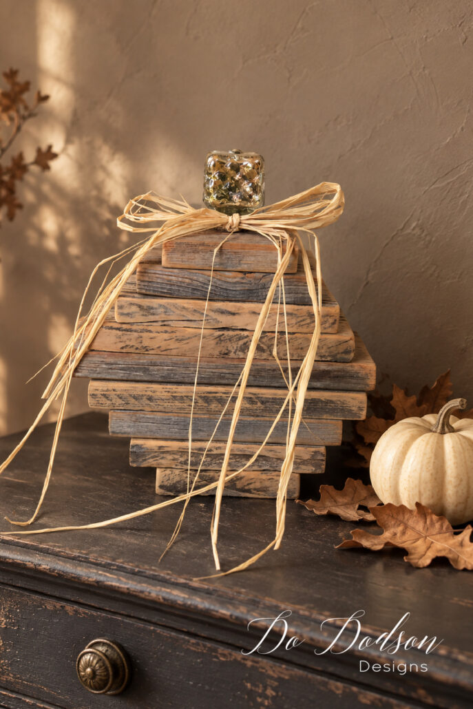 Stacked wooden pumpkin made from weathered fence pickets, topped with a glass knob and raffia bow, displayed on a dark vintage dresser with a white pumpkin and fall leaves.