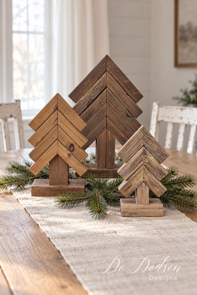Three 2x4 scrap wood Christmas trees with contrasting colorwash finishes displayed on a neutral table runner with pine greenery.