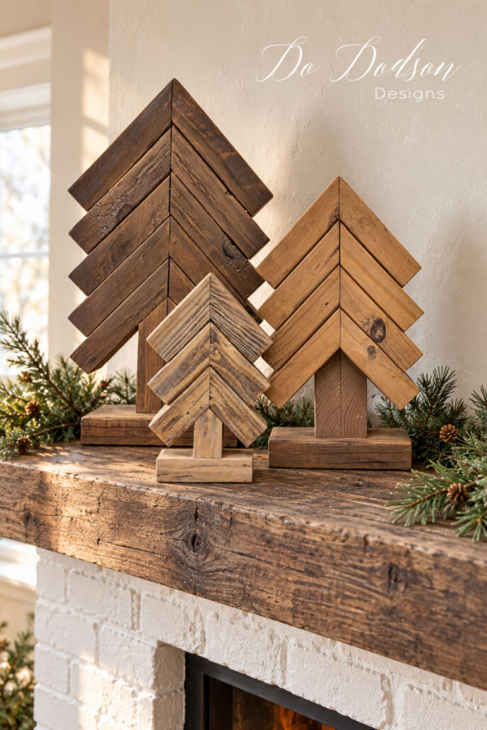 Colorwashed scrap wood Christmas tree trio arranged with pine greenery on a rustic wood mantel above a white brick fireplace.