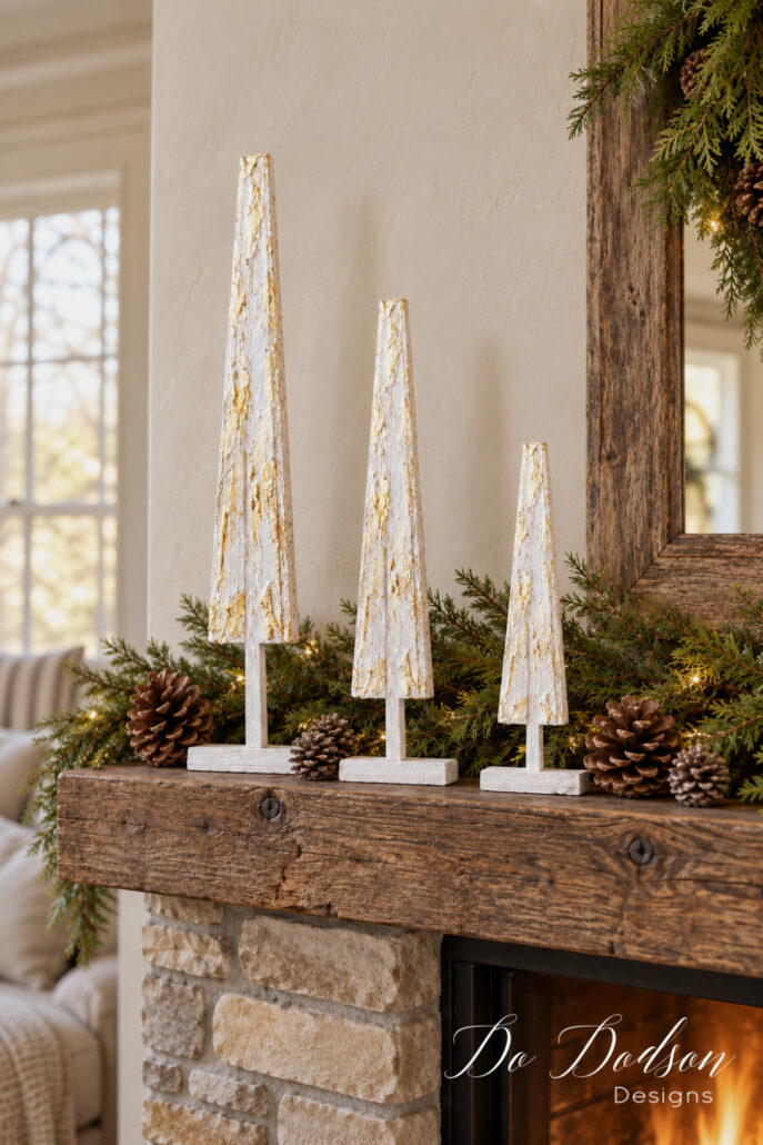 Three textured white and gold wood shim Christmas trees displayed on a rustic mantel with evergreen garland, pinecones, and warm string lights.