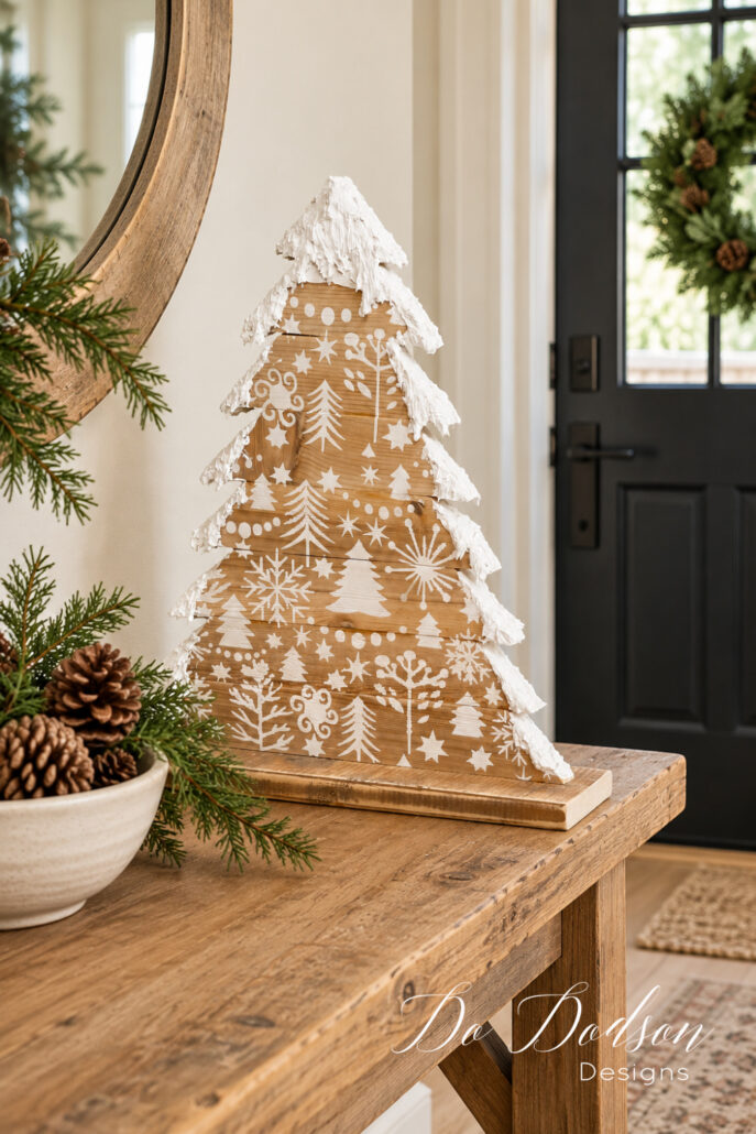 Rustic wooden Christmas tree with white stenciled designs and textured snow edges displayed on an entry table beside pinecones and greenery, with a black door behind it.