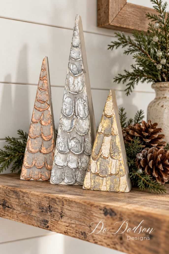 Three textured wood block Christmas trees with copper, silver, and gold accents displayed on a rustic wooden shelf with pinecones and evergreen sprigs.