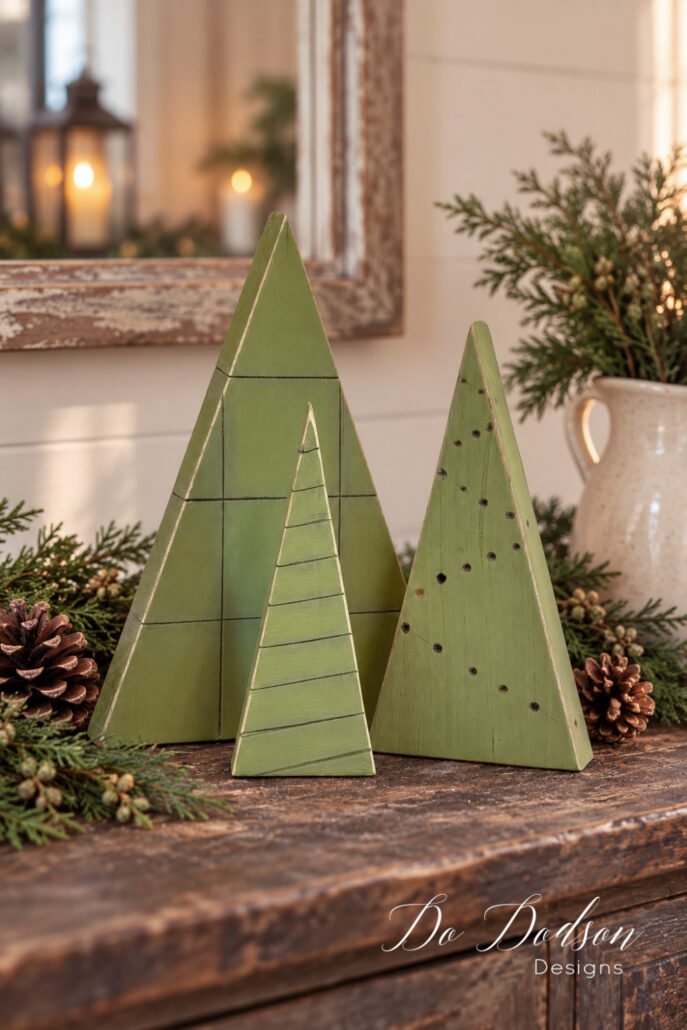 Three rustic green wood block Christmas trees with carved lines and drilled dot details displayed on a wooden table with pinecones and evergreen sprigs.