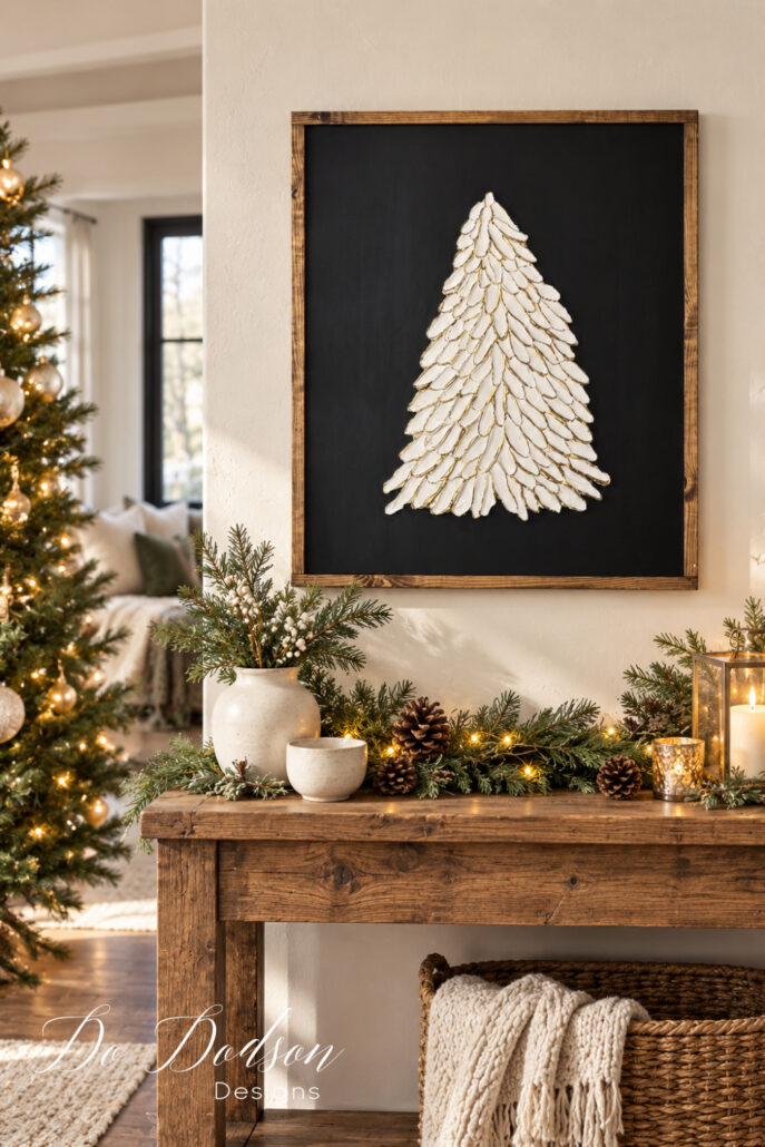 Framed white spackle Christmas tree with gold highlights on a black background, hung above a rustic console table decorated with evergreen garland, pinecones, and warm lights.